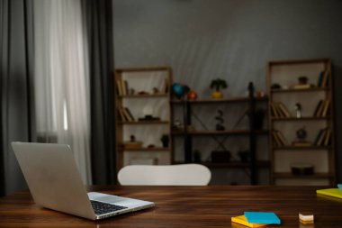 Kitap rafları ve bitkilerle çevrili ahşap bir masada açık bir dizüstü bilgisayar içeren modern bir çalışma alanı tasarımı..