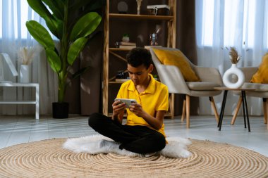 Günlük yaşam tarzı konsepti. Genç Afrikalı Amerikalı çocuk evde boş zaman geçiriyor, çocuk oturma odasında yerde oturuyor ve akıllı telefondan video oyunu oynuyor..