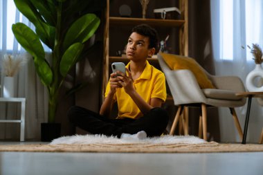 Günlük yaşam tarzı konsepti. Genç Afrikalı Amerikalı çocuk evde boş zaman geçiriyor, çocuk oturma odasında yerde oturuyor ve akıllı telefondan internete mesaj atıyor..
