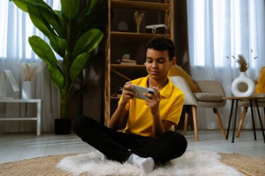 Günlük yaşam tarzı konsepti. Genç Afrikalı Amerikalı çocuk evde boş zaman geçiriyor, çocuk oturma odasında yerde oturuyor ve akıllı telefondan video oyunu oynuyor..