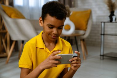 Günlük yaşam tarzı konsepti. Genç Afrikalı Amerikalı çocuk evde boş zaman geçiriyor, çocuk oturma odasında yerde oturuyor ve akıllı telefondan video oyunu oynuyor..