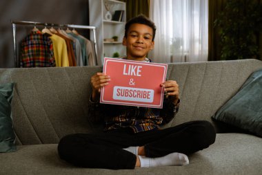 Günlük yaşam tarzı konsepti. Genç Afrikalı Amerikalı çocuk evde vakit geçiriyor, çocuk blogcu kanepede oturuyor ve yazı kağıdını beğenip abone oluyor..