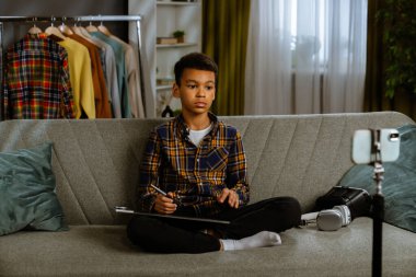 Günlük yaşam tarzı konsepti. Genç Afrikalı Amerikalı çocuk evde vakit geçiriyor, çocuk blogcu koltukta oturuyor akıllı telefondaki videoları kaydediyor, fotoğraf tableti kullanarak kameraya konuşuyor ve eleştiri yapıyor..