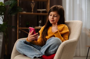 Sıradan kıyafetler giyen genç bir kız, rahat bir sandalyede rahat bir şekilde otururken akıllı telefon kullanıyor ve baş parmak işareti yapıyor..