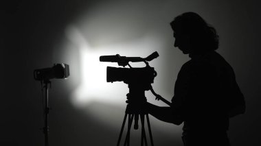 Stüdyo arkaplanına karşı üç ayaklı kamera kullanan bir kameramanın silüeti dramatik bir etki yaratıyor..