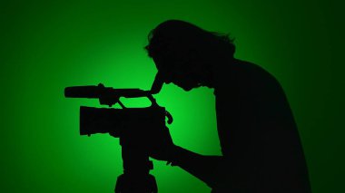 Bir kamera operatörünün silüeti canlı yeşil bir arka planda profesyonel bir kameraya bakıyor..
