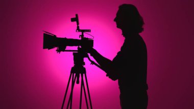 Bir insan, canlı pembe bir arkaplana karşı silueti olan üç ayaklı bir kamera kullanıyor ve sanatsal bir sahne yaratıyor..