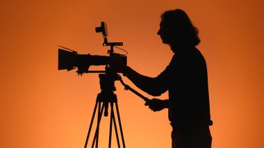 Bir kameramanın silueti üç ayaklı bir kamerayı ayarlıyor, turuncu bir zemin üzerine kurulmuş, sinematik ambiyansı yakalıyor..