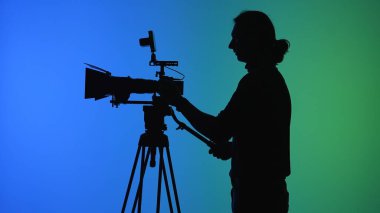 Bir kamera operatörü canlı mavi ve yeşil bir arkaplan karşısında siluetlenir, sette ekipmanları ayarlar.