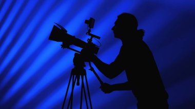 Profesyonel bir kamera arkasında çalışan bir kamera operatörünün silüeti dinamik bir mavi arka plana karşı..