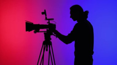 Canlı kırmızı ve mavi arka plana karşı ekipmanları ayarlayan profesyonel bir kamera operatörünün silüeti..