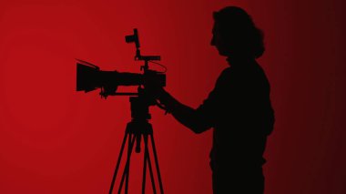 Canlı kırmızı bir arkaplana karşı tripoda monte edilmiş profesyonel bir kamera kullanan bir kişinin silüeti kontrastı vurguluyor..