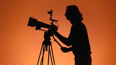Bir kameramanın silueti, kamerayı turuncu bir arkaplan önünde tripoda ayarlıyor ve film yapımının özünü yakalıyor..