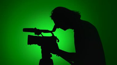 Parlak yeşil arkaplana karşı video kamera kullanan bir kişinin silueti, bir medya prodüksiyon sahnesini gözler önüne seriyor.
