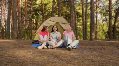 Üç kişi bir orman ortamında çadırın önünde mutlu bir şekilde oturur ve birlikte ferahlatıcı bir kamp deneyimi yaşarlar. Yavaş çekim