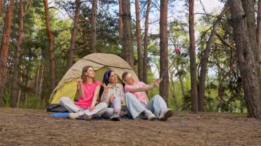 Ormandaki bir çadırın önünde oturan üç arkadaş selfie çubuğuyla anılarını yakalıyor ve dışarıda eğleniyorlar. Yavaş çekim