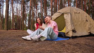 Üç arkadaş ormanda bir çadırın yanında oturmuş, yürüyüş yapıyor ve telefonda görüntülü sohbet yapıyorlar..