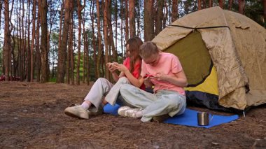 İki arkadaş çadırın yanında oturur, sohbet eder ve akıllı telefonlarıyla oyun oynarlar, ormanda yürüyüş yaparlar..