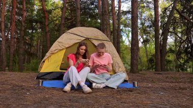 İki arkadaş çadırın yanında oturur, sohbet eder ve akıllı telefonlarıyla oyun oynarlar, ormanda yürüyüş yaparlar..