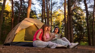 Üç arkadaş ormanda bir çadırın yanında oturmuş, bir dizüstü bilgisayarın üzerinde yürüyüş ve görüntülü sohbetin tadını çıkarıyorlar..