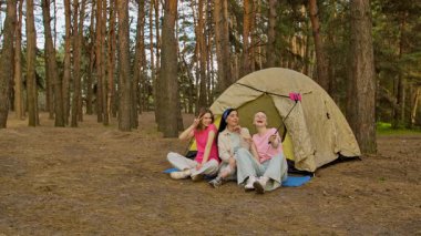 Ormandaki bir çadırın önünde oturan üç arkadaş selfie çubuğuyla anılarını yakalıyor, dışarıda eğleniyorlar..