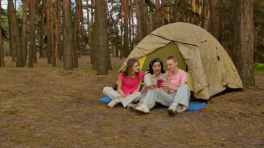 Üç arkadaş ormanda bir çadırın yanında oturmuş, yürüyüş yapıyor ve telefonda görüntülü sohbet yapıyorlar..