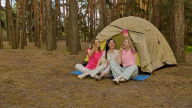Üç arkadaş ormanda bir çadırın yanında oturmuş, yürüyüş yapıyor ve telefonda görüntülü sohbet yapıyorlar..