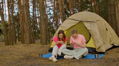 İki arkadaş çadırın yanında oturur, sohbet eder ve akıllı telefonlarıyla oyun oynarlar, ormanda yürüyüş yaparlar..