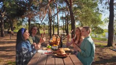 Bir grup park ortamında pikniğin tadını çıkarıyor, sıcak bir atmosferde ızgara sosis ve taze yiyecek paylaşıyorlar. Yavaş çekim