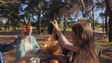 Bir grup parkta piknik yapıyor, bira içiyor, sıcak bir atmosferde ızgara sosis ve taze yiyecek paylaşıyor. Yavaş çekim