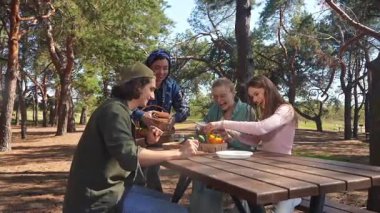 Bir grup arkadaş güneşli bir ormanda piknik yapıyorlar, sohbet ediyorlar ve açık havada taze sebzelerin tadını çıkarıyorlar..