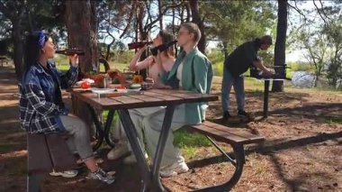 Orman ortamında piknik yapan bir grup arkadaş, ahşap bir masada kahkaha ve yemek paylaşıyorlar..