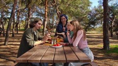 Bir grup arkadaş güneşli bir park ortamında piknik masasında dinleniyorlar, birbirlerine eşlik ediyorlar ve dışarıda eğleniyorlar..