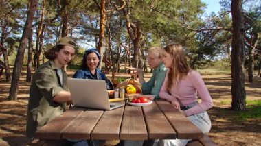 Bir grup arkadaş parkta bir piknik masasında yemeğin tadını çıkarırken bir yandan da bilgisayardan video görüşmesi yapıyorlar..