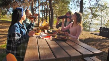 Orman ortamında piknik yapan bir grup arkadaş, ahşap bir masada kahkaha ve yemek paylaşıyorlar..