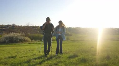 İki yürüyüşçü çimenli bir arazide duruyor, güneşli havanın ve uzaktaki resimli manzaranın tadını çıkarıyorlar. Yavaş çekim