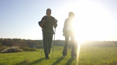 İki yürüyüşçü çimenli bir arazide duruyor, güneşli havanın ve uzaktaki resimli manzaranın tadını çıkarıyorlar. Yavaş çekim