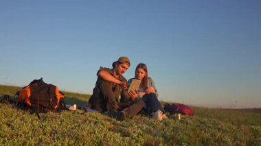 Sırt çantalarıyla çimenlerde oturan genç bir adam ve kadın açık mavi gökyüzünün altında tablet kullanıyorlar. Yavaş çekim