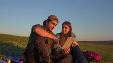 Sırt çantalarıyla çimenlerde oturan genç bir adam ve kadın açık mavi gökyüzünün altında tablet kullanıyorlar. Yavaş çekim