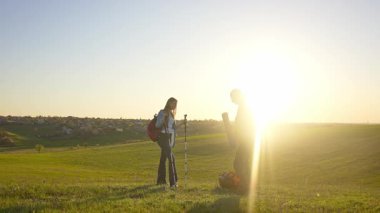 İki yürüyüşçü çimenli bir arazide durur, güneşli havanın ve uzaktaki manzaranın tadını çıkarırlar..