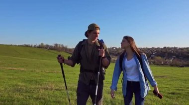 Açık mavi gökyüzünün altında çimenli bir arazide yürüyüş yapan bir çift, somut bir macera ve açık hava keşfi..