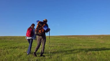 İki yürüyüşçü çimenli bir arazide durur, güneşli havanın ve uzaktaki manzaranın tadını çıkarırlar..