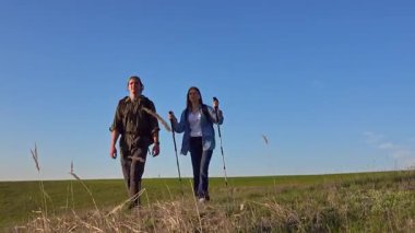 Açık mavi gökyüzünün altında çimenli bir arazide yürüyüş yapan bir çift, somut bir macera ve açık hava keşfi..