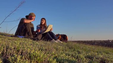 Genç bir çift çimenli bir tepede oturmuş haritayı inceliyor. Açık mavi gökyüzünün altında bir açık hava macerasına hazırlanıyorlar..