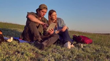 Sırt çantalarıyla çimenlerde oturan genç bir adam ve kadın açık mavi gökyüzünün altında tablet kullanıyorlar..