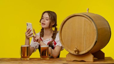 Geleneksel giyinmiş genç bir kadın iki bira bardağı ve ahşap bir fıçının yanında oturuyor, akıllı telefonunu parlak sarı bir arka plana karşı kullanıyor..