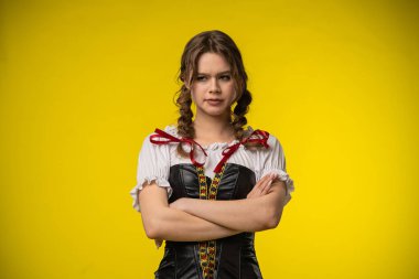 Geleneksel Bavyera elbisesi giymiş genç bir kadın kollarını kavuşturup parlak sarı bir arkaplanda poz veriyor..