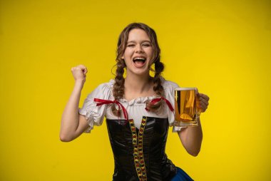 Geleneksel Bavyera kıyafetli neşeli genç bir kadın elinde bira bardağıyla sarı bir arka planda kutlama yapıyor..