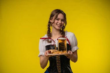 Geleneksel giyinmiş bir kadın, elinde bira ve krakerle canlı sarı bir zemine gülümser..