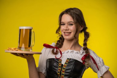 Geleneksel giysiler içinde gülümseyen bir kadın, büyük bir bira bardağını ve krakerleri canlı sarı bir arka plana karşı bir tepside tutarak neşeli ve neşeli bir atmosferi çağrıştırıyor..